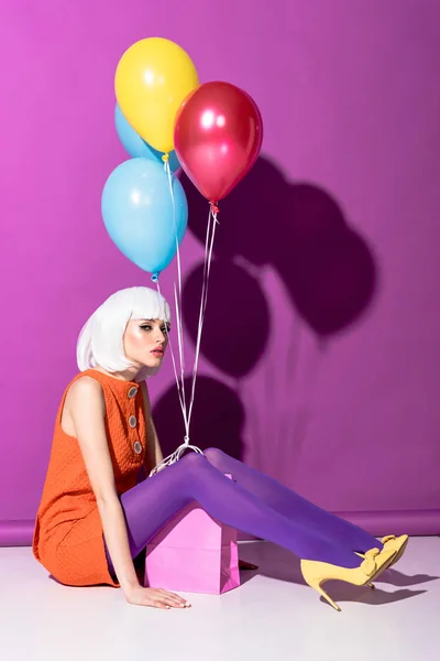 Serious girl in white wig with air balloons sitting on purple background — Stock Photo