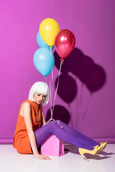 Sad young woman in white wig with air balloons sitting on purple background — Stock Photo