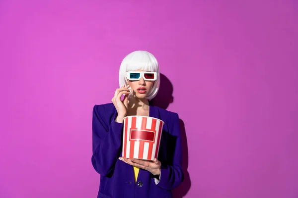 Mujer sorprendida en peluca y gafas 3d comiendo palomitas de maíz sobre fondo púrpura - foto de stock