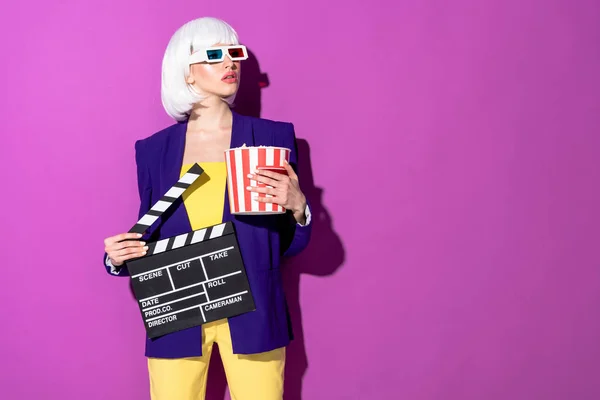 Interested girl in 3d glasses holding clapperboard and popcorn and looking away on purple background — Stock Photo