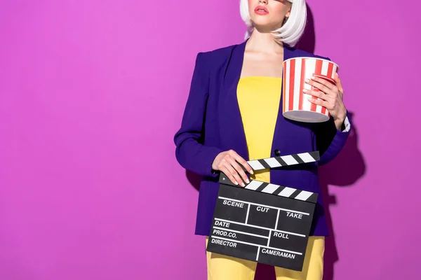 Cropped view of woman in blue jacket holding popcorn and clapperboard on purple background — Stock Photo