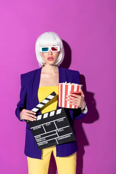 Amazed girl in 3d glasses holding popcorn and clapperboard on purple background — Stock Photo