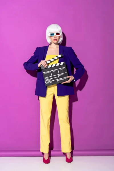 Stylish young woman in 3d glasses holding clapperboard on purple background — Stock Photo