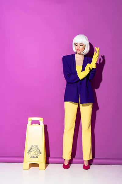 Girl in wig standing near wet floor sign and putting on rubber gloves on purple background — Stock Photo