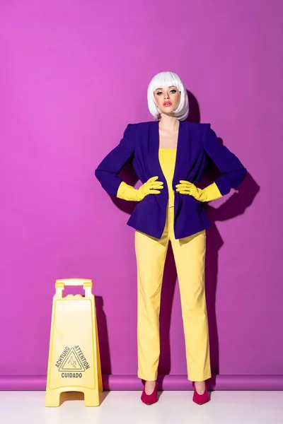 Muchacha en peluca y guantes de goma de pie con brazos akimbo cerca del cartel del piso mojado sobre fondo púrpura - foto de stock