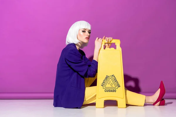 Chica glamurosa en peluca sentada en el suelo con cartel de suelo húmedo sobre fondo púrpura — Stock Photo