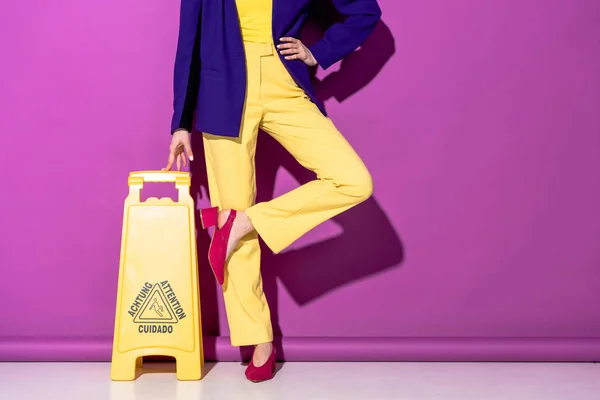 Cropped view of stylish woman standing on one leg near wet floor sign on purple background — Stock Photo