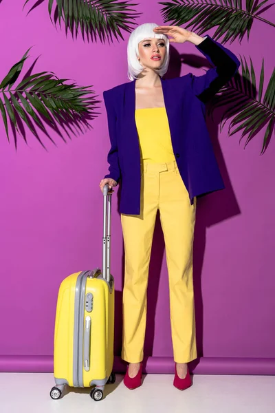 Стильная женщина в парике с чемоданом, смотрящая на фиолетовый фон — стоковое фото