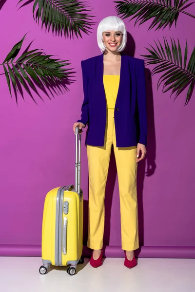 Visão de comprimento total da menina sorridente na peruca com mala amarela em pé sobre fundo roxo — Fotografia de Stock