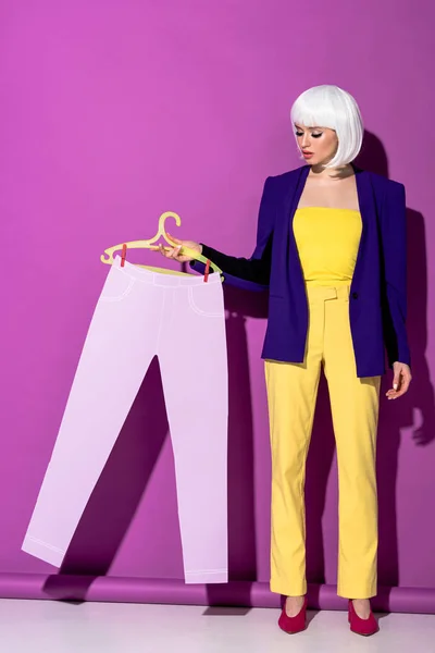Вид на повну довжину елегантної дівчини в білій перуці, що тримає вішалку з паперовими штанами на фіолетовому фоні — стокове фото