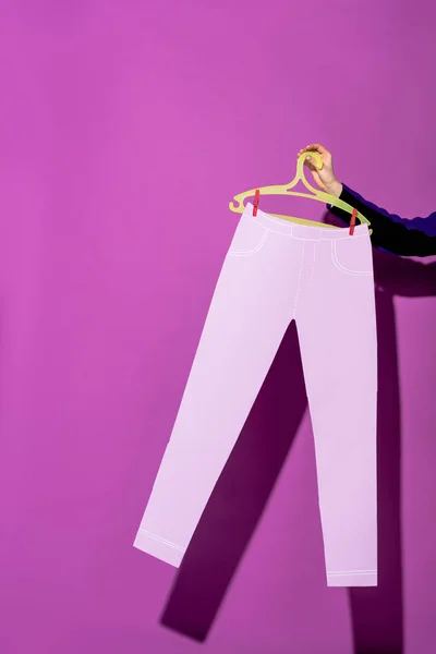 Vista cortada de mulher segurando cabide com calças de papel no fundo roxo — Fotografia de Stock