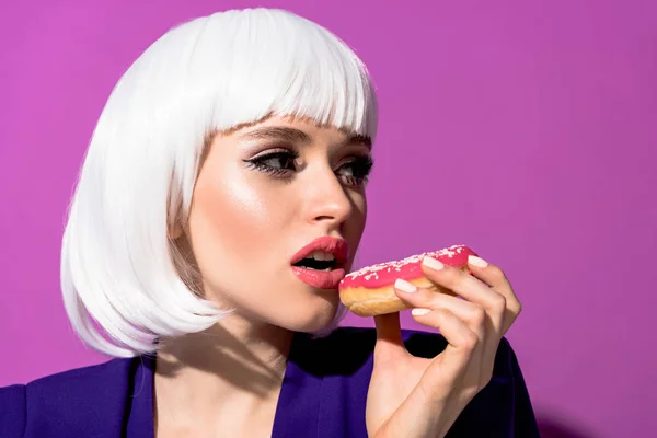 Linda chica en peluca blanca comiendo donut sobre fondo púrpura - foto de stock