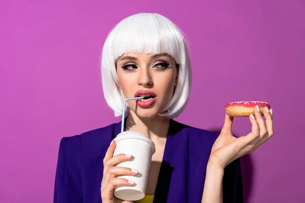 Girl in wig drinking beverage and holding doughnut on purple background — Stock Photo
