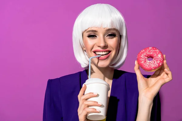 Mulher sorridente na peruca bebendo bebida e segurando donut no fundo roxo — Fotografia de Stock