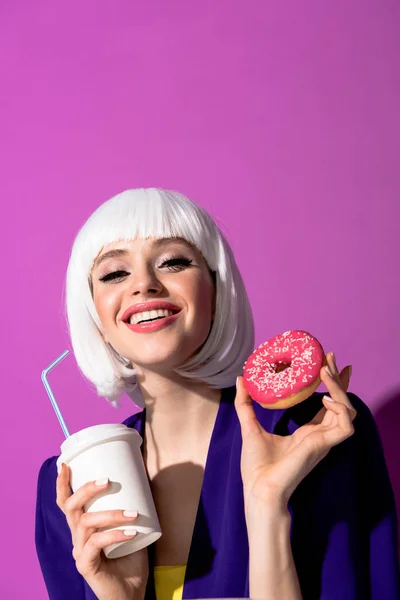 Весела дівчина в перуці тримає напій і пончик на фіолетовому фоні — стокове фото