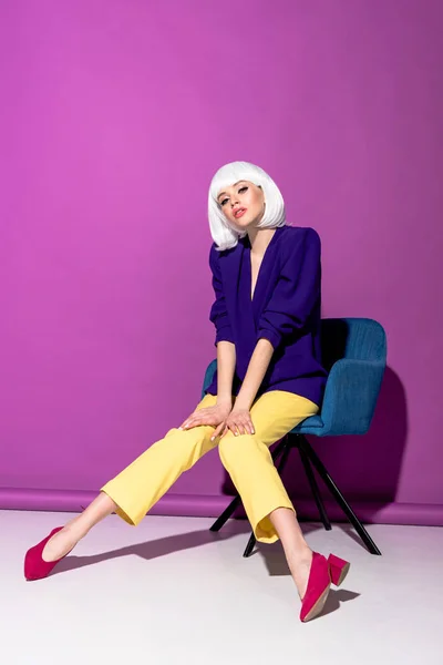 Attractive girl in white wig sitting in armchair on purple background — Stock Photo