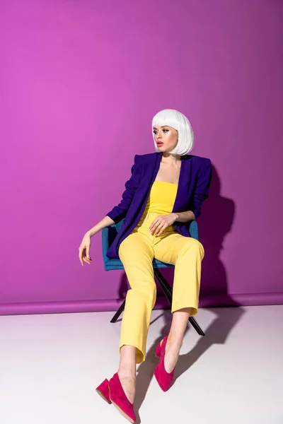 Elegante chica de peluca blanca sentada en sillón y mirando hacia otro lado sobre fondo morado - foto de stock