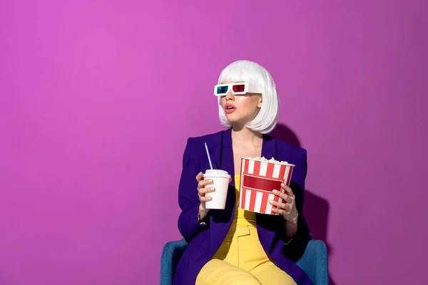 Femme étonnante dans des lunettes 3D assis dans un fauteuil avec soda et pop-corn sur fond violet — Photo de stock