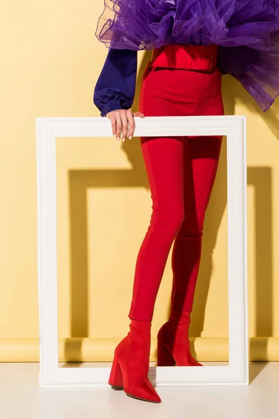 Vue recadrée de la femme en pantalon rouge tenant le cadre blanc sur fond jaune — Photo de stock