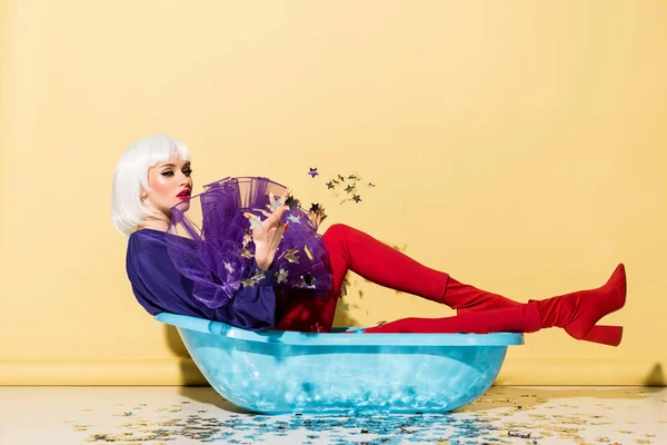 Attractive girl in wig lying in blue tub and throwing out star confetti on yellow background — Stock Photo