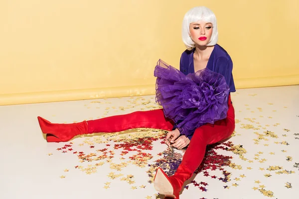 Pensive woman in wig and red pants sitting on star confetti on yellow background — Stock Photo