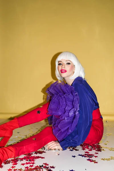 Smiling woman in white wig sitting on confetti on yellow background — Stock Photo