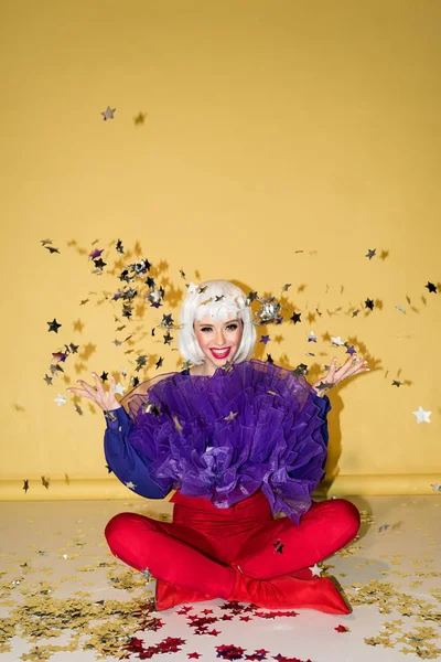 Счастливая девушка в белом парике сидит на полу со скрещенными ногами и выбрасывает звездные конфетти на желтый фон — стоковое фото