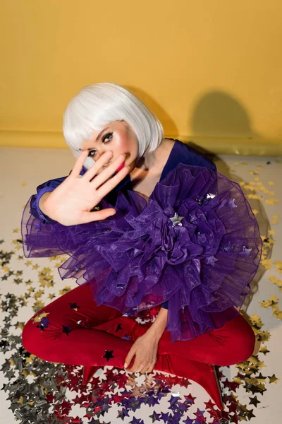 Woman in wig sitting on confetti with crossed legs and outstretched hand on yellow background — Stock Photo