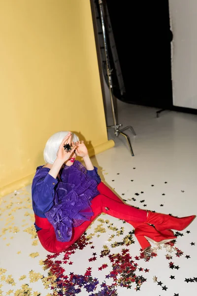 Woman in wig sitting on confetti with crossed legs and covering face with hands on yellow background — Stock Photo