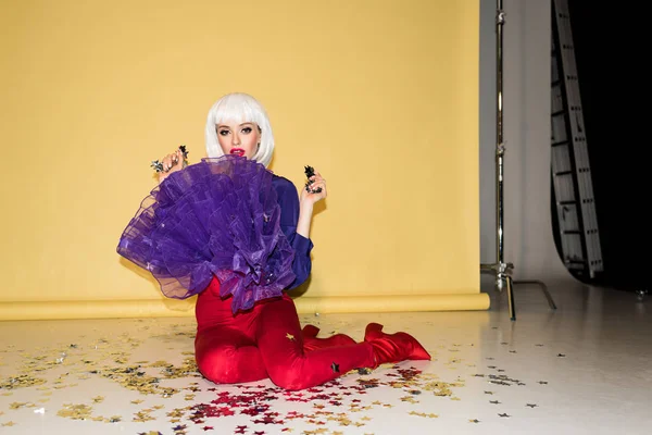 Gorgeous girl in wig sitting on floor and holding confetti on yellow background — Stock Photo