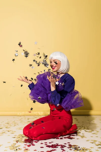 Chica alegre en peluca blanca lanzando confeti estrella sobre fondo amarillo - foto de stock