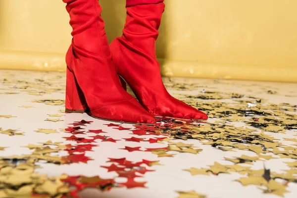 Cropped view of woman in red high-heeled shoes standing on confetti on yellow background — Stock Photo