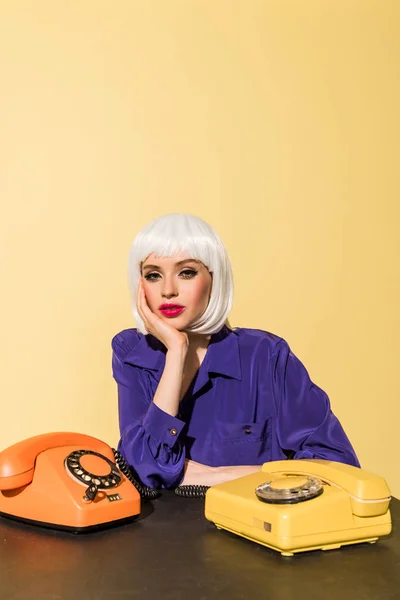 Mujer curiosa en peluca posando con teléfonos viejos aislados en amarillo - foto de stock