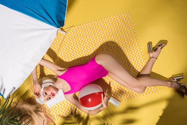 Vista superior da menina em maiô rosa e sapatos de salto alto deitado com bola na toalha amarela — Fotografia de Stock