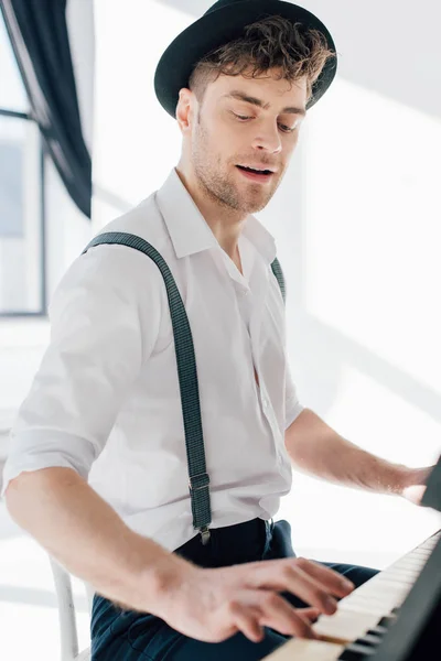 Enfoque selectivo de pianista con estilo en camisa blanca y sombrero negro tocando el piano en casa - foto de stock