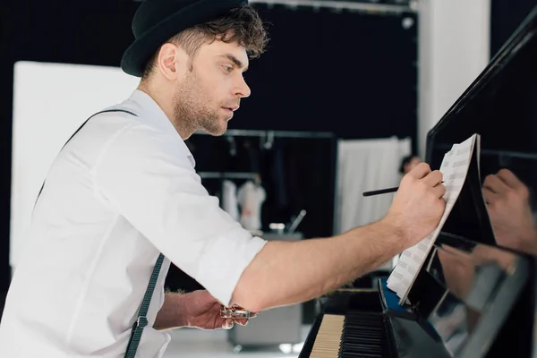 Selective focus of handsome composer writing in music book while sitting at piano at home — Stock Photo