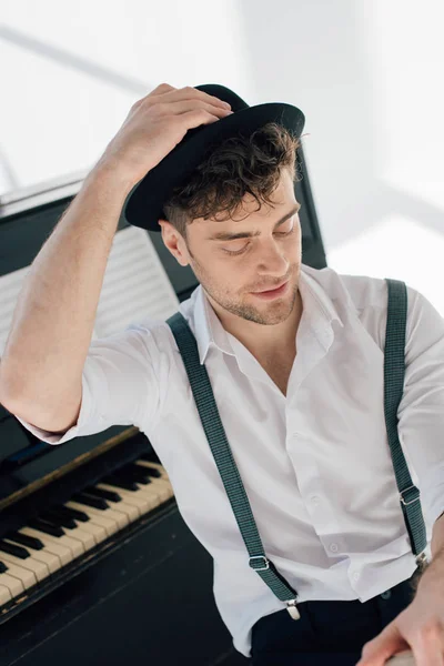 Pianiste souriant dans des vêtements branchés assis au piano et tenant la main sur le chapeau — Photo de stock