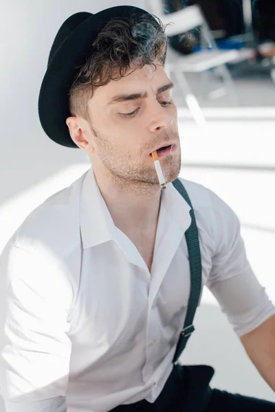 Hombre serio en camisa blanca y sombrero negro fumar cigarrillo - foto de stock