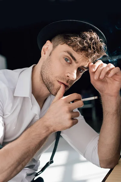 Hombre guapo pensativo en camisa blanca y sombrero negro fumar cigarrillo - foto de stock