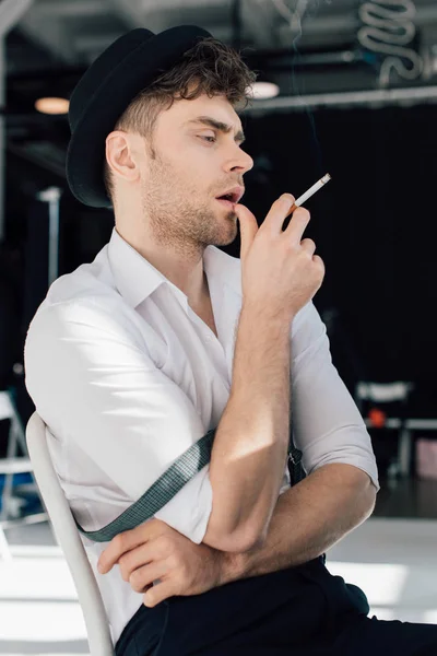 Bel homme rêveur en chemise blanche et chapeau noir fumant cigarette — Photo de stock