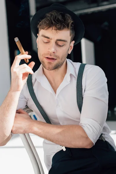 Pensive man in white shirt and black hat holding cigar while sitting on chair — Stock Photo