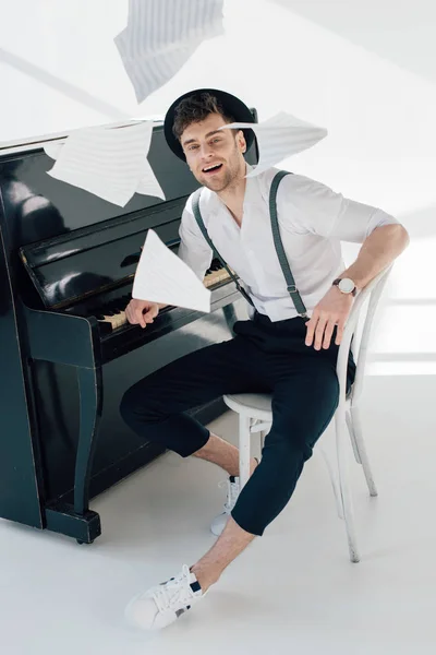 Smiling musician in trendy clothing sitting by piano and throwing music book sheets — Stock Photo