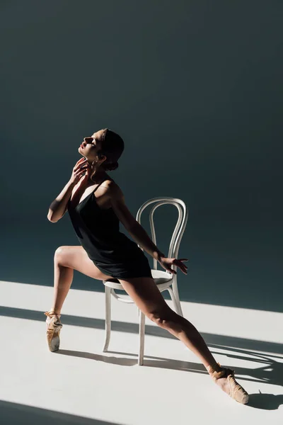 Joven bailarina en vestido negro estiramiento en silla blanca en la luz del sol - foto de stock