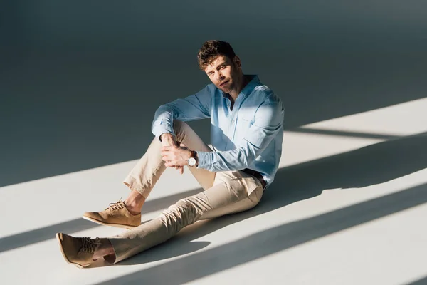 Handsome man in blue shirt and beige jeans sitting in sunlight and looking at camera — Stock Photo