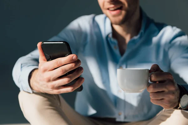 Messa a fuoco selettiva dell'uomo utilizzando smartphone e tenendo tazza di caffè — Foto stock