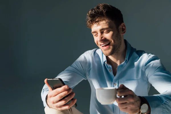 Hombre alegre usando teléfono inteligente y sosteniendo la taza de café - foto de stock