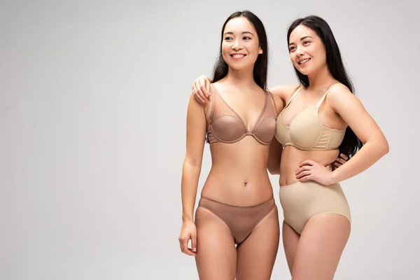 Zwei hübsche multiethnische Frauen in Dessous, die sich umarmen und isoliert auf grauem, körperpositivem Konzept lächeln — Stockfoto