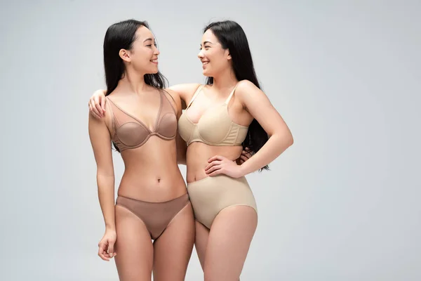 Zwei fröhliche multiethnische Frauen in Dessous, die sich umarmen und einander isoliert auf grauem, körperpositivem Konzept betrachten — Stockfoto