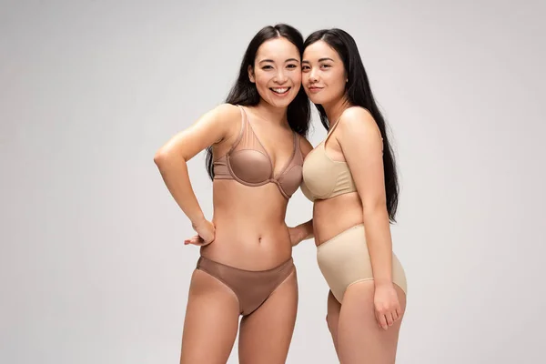 Zwei glückliche multikulturelle Mädchen in Unterwäsche, die sich umarmen und in die Kamera schauen, isoliert auf grauem, körperpositivem Konzept — Stockfoto