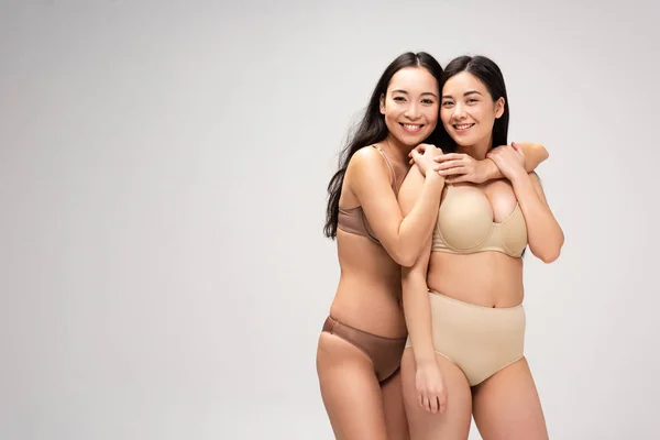 Две весёлые мультикультурные девушки в нижнем белье обнимаются и смотрят на камеру, изолированную на сером, концепция позитивности тела — стоковое фото